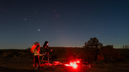 Person using a telescope in a desert landscape under a starry night sky.