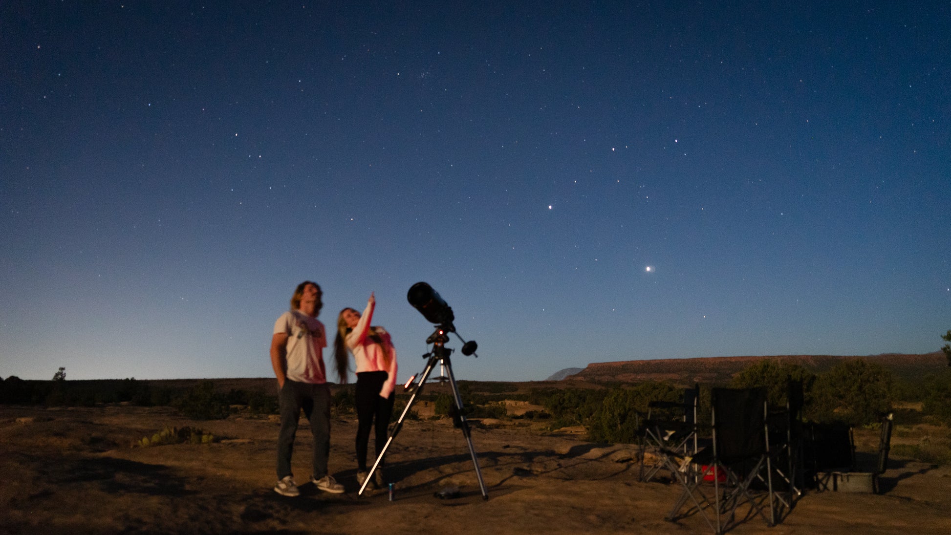 Two people with a telescope under a starry night sky in a desert landscape.