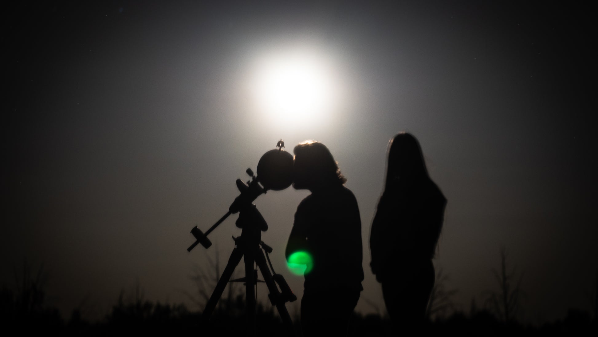 Two people silhouetted against the moon, one holding a telescope.