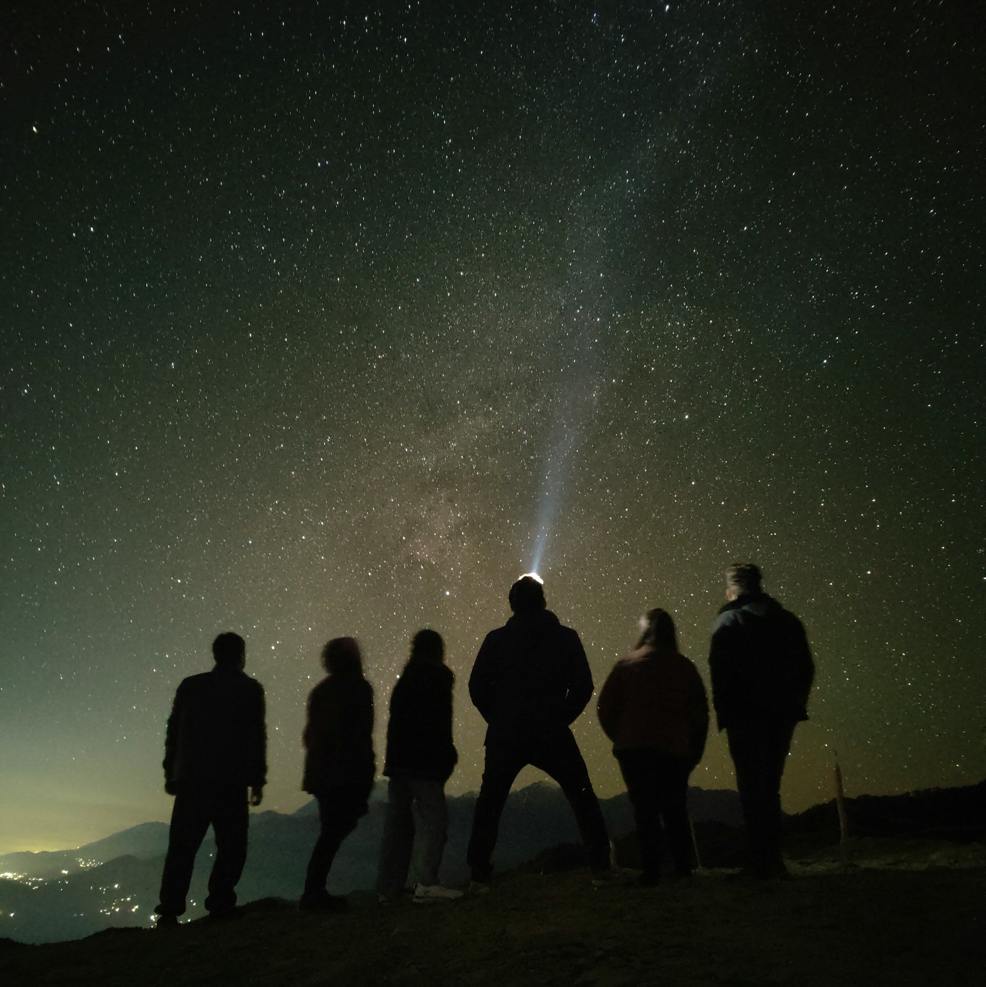 Silhouettes of people on a mountain top under a starry night sky, with one person holding a flashlight.
